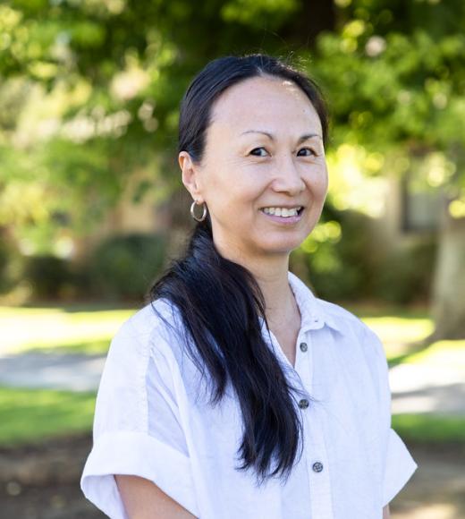 A woman with long black hair smiles outdoors, surrounded by green trees and grass.